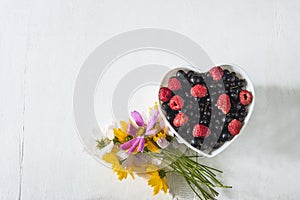 Red raspberries and red currants in the white bowl shape heart and bouquet of wild flowers