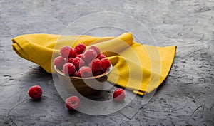 Red raspberries placed in a round wooden bowl accompanied by a yellow napkin, all on a gray background