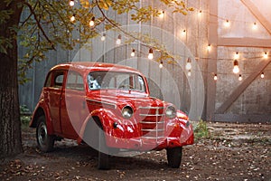 Red retro car standing in the garden in the summer on a background of gray wall and burning bulbs. Vintage vehicle near the garage