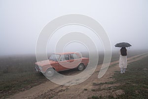 Red retro car stands near the dirt road in fog, young girl in white coat stands under umbrella