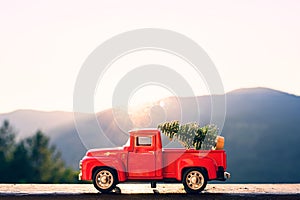 Red retro small car carrying christmas tree in the mountain in the sunbeams of sunrise. Winter holiday concept.