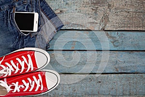 Red retro sneakers and smartphone in a jeans pocket on a blue wooden background