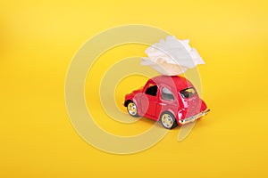 Red retro toy car with white rose flower on yellow background. Flowers, gifts delivery concept.