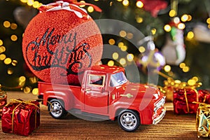 Red retro toy truck with Christmas ball and gift boxes on wooden table over background with christmas lights. Greeting card Marry