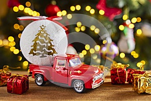 Red retro toy truck with Christmas ball and gift boxes on wooden table over background with christmas lights. Greeting card Marry