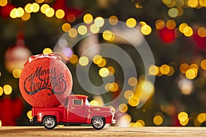 Red retro toy truck with Christmas ball on wooden table over background with christmas lights. Greeting card Marry Christmas and
