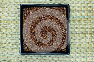 Red Rice Grains in Black Plate on Natural Bamboo Mat Background Surface with Free Space