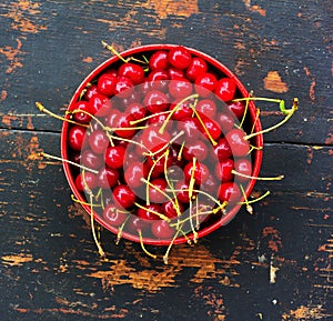 Red ripe cherries with tails in a circular plate on an old black wooden background with a crack
