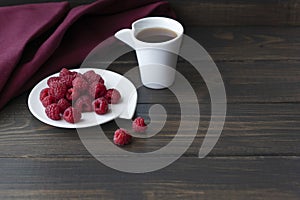 red ripe raspberries are on a white plate, next to them there is a cup of vitamin tea and red textile on wooden background, copysp