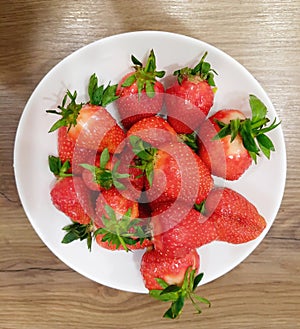 Red ripe strawberries on a white plate on the brown surface of the table.