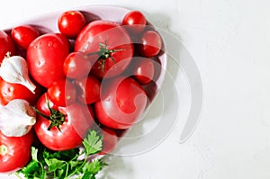 Red ripe tomatoes of different sizes with garlic and a sprig of parsley on a pink large plate on a white background.