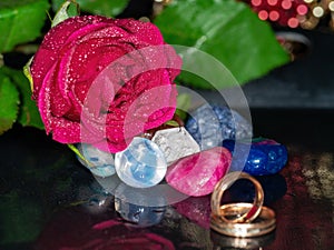 Red rose covered in water beads colorful gem stone in focus, Wedding rings in foreground out of focus. Concept marriage, romance