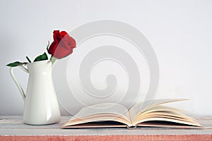 A red rose flower in a white vintage vase beside an open book.