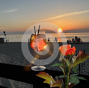 red roses and glass of orange  water  with ice  on wooden table top    sunset at sea  in beach restaurant view in pink sky and se