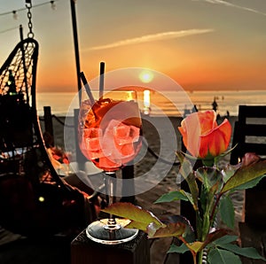 Red roses and glass of wine  with ice  on wooden table top    sunset at sea  in beach restaurant view in pink sky and se