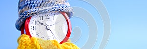 Red round alarm clock in knitted wool blue hat and yellow scarf on a blue background. winter time concept. winter season. cozy and