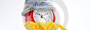 Red round alarm clock in knitted wool blue hat and yellow scarf on a white background. winter time concept. winter season. cozy an