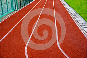 Red running track with white lines in outdoor sport stadium, side is a field and park. Backgrounds and rubber texture