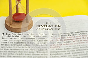 Red sand hourglass on top of open Holy Bible in the Book of Revelation of Jesus Christ chapter 1.
