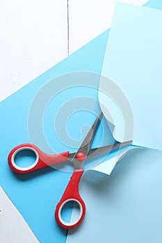 Red scissors cutting light blue paper on white wooden background, top view