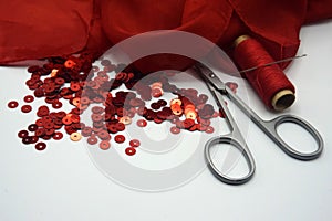 Red sequins, spool of red thread,needle and scissors on rippled red silk background.