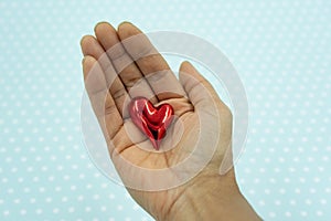 Red shiny heart in the palm of mixed race woman at white dotted blue background.
