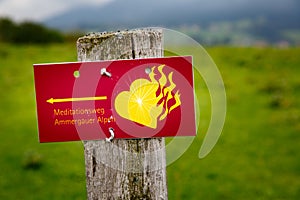 Red sign in German: Ammergau Alps meditation trail. With a yellow flaming heart