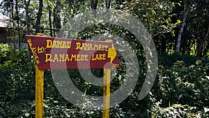 Red sign next to green vegetation giving easy direction with yellow letters and arrow towards ranamese lake. photo