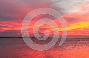 Red sky and ocean near end of stunning sunset with purple and yellow streaks and cars going over bridge and lights of shore in dis