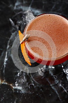 A red sour cocktail in a coupe glass garnished with an orange peel. Clover club with imported gin.