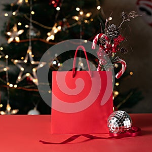 Red sparkling shopping bag mockup with decorative pine tree branch, candy canes, mirror ball on red table with blurred
