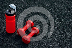 Red sports water bottle and sports dumbbells on a black rubberized sports field, background image