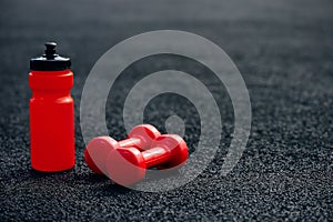 Red sports water bottle and sports dumbbells on a black rubberized sports field in the background light, background image