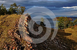 Red stone rocks in yellow dry grass on bay of lake baikal in autumn. Blue water, clouds, trees. Sunset