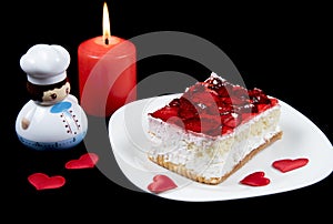 Red strawberry cake on white plate, candle, hearts and toy cook isolated on black background.  Holiday food for Valentine day,