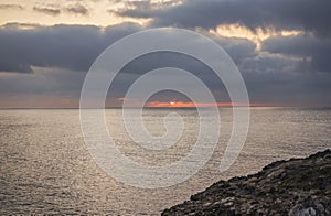 Red sun rising above sea behind clouds, rocky shore near