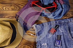 Red sunglasses, straw hat, summer scarf, jeans shorts and nail polish on wooden background