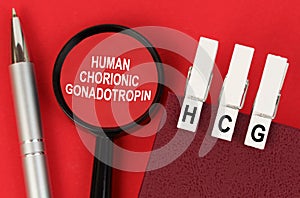 On the red surface lies a pen, a notebook with clothespins - HCG, and a magnifying glass - human chorionic gonadotropin