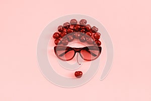 Red sweet cherry laid out in image of woman in sunglasses with lips on pink background, coral toned. Concept youth, beauty,