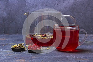 Red tea rooibos, hibiscus, karkade in glass cup and teapot among dry tea leaf, petals and cranberries on a dark background.