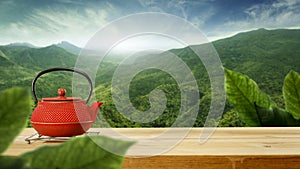 A red teapot on a metal stand on wooden table with beautiful green trees mountains and blue cloudy sky background.