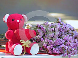 Red teddy bear sit holding a red heart with gold ribbon with bouquet of flowers lying on the table