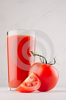 Red tomato juice in glass and juicy slice cut tomato on soft white wood board, closeup, vertical.