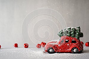 Red toy car with a Christmas tree on the roof in the snow, on a gray background with red New Year`s toys. Place for text