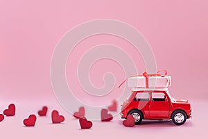 Red toy car delivering carrying on roof gift box with small red hearts on pink background. Valentine`s day concept