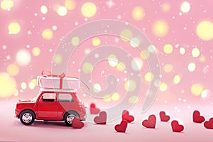 Red toy car delivering carrying on roof gift box with small red hearts on pink background. Valentine`s day concept