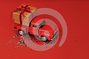 Red toy truck with craft gift box and red bow in its trunk. Miniature car with Christmas decoration on red bright background with