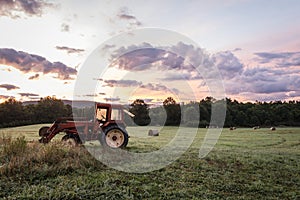 Red tractor and freshly rolled hay bales rest on rolling hill with dramatic cloudscape at sunrise