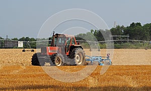 Red tractor with plough machine plows a just cut hay field. Large water irrigation system on background