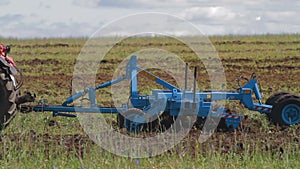 Red tractor with a reverse plow plows a field. A stork walks nearby and collects food. Agricultural work in the field